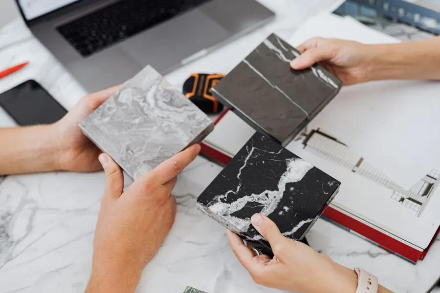 Two people comparing marble and stone tile samples on a desk beside a laptop