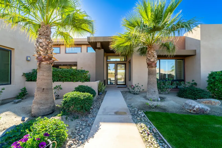 The front walk of a finished stucco home between two palms and desert landscaping, leading to a glass entry door