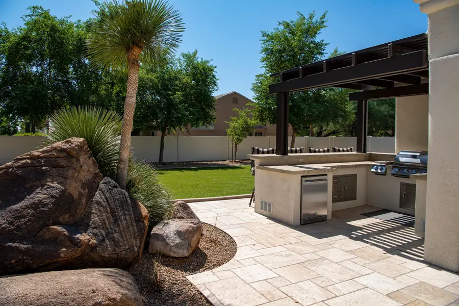 An outdoor kitchen with a built-in grill and bar under a dark wood pergola, beside a travertine patio and desert boulders